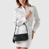 Cartera tipo baguette de la marca Guess, color negro con textura granulada. Presenta un logo triangular metálico en el frente y un charm colgante en forma de corazón con borla. Incluye una correa ajustable para el hombro y cierre superior.