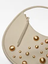 Cartera hobo redonda color crema, con asa de hombro y cierre de cremallera. Decorada con tachas doradas de diferentes tamaños en el frente.