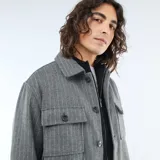 Sobrecamisa de corte recto y diseño estructurado, confeccionada en tejido gris con patrón de rayas verticales finas. Presenta cuello clásico, cierre frontal con botones y dos bolsillos frontales con solapa.