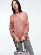 Camisa de corte oversize confeccionada en tejido jersey con acabado brillante. Presenta cuello en punta, cierre frontal con botones, mangas largas con puños anchos y tajos laterales en el ruedo.