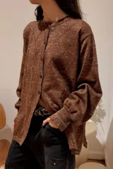 Camisa de punto con textura brillante, color marrón, de corte holgado, cuello redondo y cierre frontal con botones. Presenta un diseño de rombos calados y mangas largas con puños ajustados.