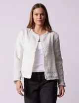 Chaqueta de tejido tweed color blanco con destellos brillantes, de corte recto y estructurado. Presenta cuello a la base, abertura frontal y puños decorados con botones en contraste. Los bordes del cuello, la cartera frontal y el ruedo cuentan con una terminación deshilachada.