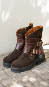 Botas de caña media combinadas en gamuza y cuero vacuno color marrón oscuro. Presentan un diseño con tiras decorativas cruzadas sobre el empeine con hebillas metálicas, cierre lateral y suela de goma con plataforma dentada.