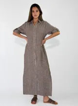 Vestido largo camisero color marrón, de corte recto y suelto, confeccionado en lino. Cuenta con mangas tres cuartos con tirilla y botón para ajustar, cuello camisero y cierre frontal completo con botones. Presenta detalles de costuras decorativas blancas a lo largo de la botonera.