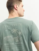 Remera verde con estampado en la espalda de una playa con palmeras y la frase "Ride the waves".