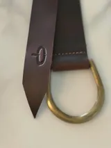Cinto de cuero flexible y envolvente con diseño minimalista, que se anuda mediante una hebilla metálica en forma de D. El extremo presenta el logo de la marca grabado.