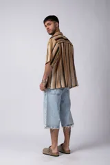 Camisa de manga corta con cuello solapa y cierre frontal de botones. Presenta un diseño de rayas verticales en tonos marrones, beige y negro, con una textura tejida y corte holgado.