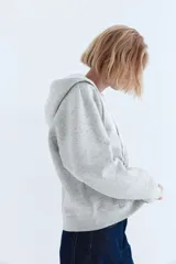 Campera con capucha y cierre frontal, color gris melange, confeccionada en felpa con interior cepillado suave. Presenta hombros caídos, bolsillos canguro y puños y bajo en punto acanalado.