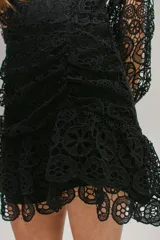 Vestido corto de encaje negro con diseño calado, escote en V, mangas largas con puños y falda con volados. Presenta un corte ajustado con detalle fruncido en la cintura y cierre invisible.