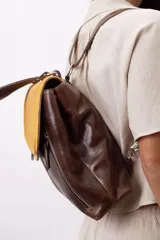 Mochila de cuero vacuno marrón con acabado pulido, solapa en forma de V y costuras decorativas. Cuenta con cremalleras laterales y asas ajustables.