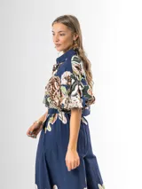 Conjunto de dos piezas confeccionado en viscosa con estampado floral en tonos marrones y verdes sobre base blanca. Incluye una blusa tipo camisa corta con cuello solapa, cierre frontal de botones y mangas abullonadas con diseño globo, y un pantalón tipo culotte de tiro alto, amplio y fluido con pinzas en la cintura.