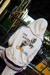 Campera con capucha de color crema, con diseño estampado en la espalda que incluye un caballo y la frase The Winter Carousel. Presenta bandas laterales con inscripciones en las mangas y un borde inferior con franjas horizontales en tonos bordó y celeste.