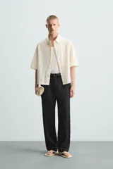 Camisa de corte relajado (relaxed fit) color crema o blanco roto, confeccionada en tejido con mezcla de algodón. Presenta un bordado sutil en relieve con patrón de ondas o líneas diagonales entrecruzadas. Tiene cuello solapa, manga corta y un bolsillo de plastrón en el pecho.