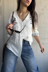 Camisa blanca de manga larga con escote en V y cierre frontal de botones. Presenta delicados bordados florales en tono oscuro en los hombros y puños.