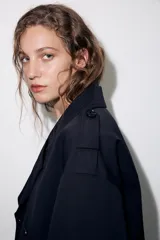 Trench corta de color negro con cuello de solapa, cierre frontal cruzado con botones y mangas largas con trabillas ajustables. Cuenta con bolsillos delanteros de vivo.
