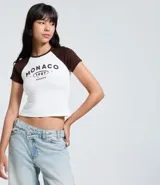 Remera ajustada de algodón con mangas cortas estilo raglán en color marrón oscuro y cuerpo blanco. Presenta un estampado central con el texto "MONACO 1987 EUROPE" en color marrón.