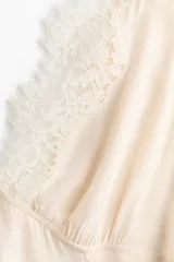 Vestido midi color beige de viscosa, sin mangas, con escote en V profundo adelante y atrás decorado con ribetes de encaje. Presenta una falda acampanada con bajo asimétrico y costuras en zigzag.