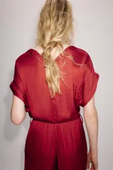 Mono largo satinado de color rojo, con escote en V cruzado y manga corta. Presenta cintura elástica con cinturón negro incorporado, bolsillos laterales ocultos y terminación de las piernas con puño elástico.