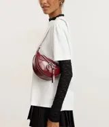 Cartera tipo riñonera de tamaño pequeño, confeccionada en poliuretano con acabado brillante. Presenta un diseño de media luna con cierre de cremallera metálica, decorado con tiradores circulares. Incluye una correa de cadena metálica para llevar al hombro o cruzada.