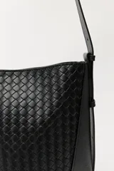 Cartera de hombro color negro con diseño trenzado en el panel frontal y laterales lisos. Cuenta con una correa larga ajustable y una placa metálica pequeña con el logo de la marca en la parte inferior.