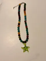 Collar de piedras naturales en tonos vibrantes como verde, azul, naranja y bordó, con un dije de estrella de mar de cristal verde con lunares blancos. Cuenta con cierre de mosquetón y cadena de extensión plateados.