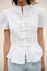 Camisa blanca de manga corta abullonada, con cuello mao y cierre frontal con alamares. Presenta corte evasé con detalle de volados en el bajo.