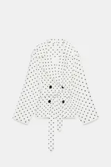 Cazadora estilo trench corta confeccionada en lino, con diseño de lunares negros sobre fondo blanco. Cuenta con cuello solapa, manga larga con trabillas en los puños, cierre frontal cruzado con botones negros y cinturón ajustable del mismo tejido.