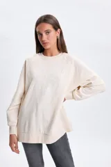 Buzo deportivo de corte oversize en color crudo, con cuello redondo y mangas largas. Presenta un diseño bordado en el frente con la inscripción Saint Tropez y un motivo floral central.