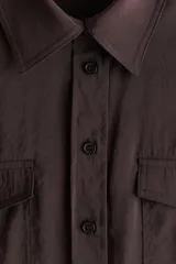 Camisa marrón de manga larga, corte corto y diseño holgado, con cuello clásico, cierre frontal de botones, bolsillos con solapa en el pecho y cordón ajustable en el bajo.