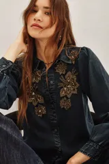 Camisa de liocel color azul oscuro, con cuello clásico y cierre frontal de botones. Presenta mangas largas abullonadas con puños elásticos y un diseño floral bordado con cuentas doradas y lentejuelas en el frente y los hombros.