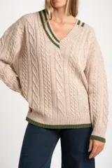 Sweater de lana y nylon color beige jaspeado, con diseño de punto trenzado (cable knit) en todo el cuerpo y mangas. Presenta cuello en V con ribete de dos franjas verdes y puños y bajo con ribete verde liso.