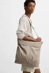 Bolso tipo shopper de tejido técnico color beige, con dos asas de mano y una correa de hombro ajustable y extraíble. Presenta un compartimento principal con cierre de cremallera, un bolsillo interior con cremallera y dos bolsillos frontales con cierre de botón a presión.