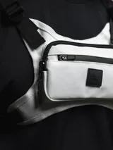 Bolso de pecho táctico color gris claro con detalles en negro, con correas ajustables y hombreras de neopreno. Cuenta con tres compartimentos: uno trasero de red y dos frontales con cierre.