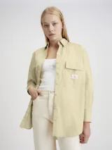 Camisa oversized color beige, confeccionada en algodón regenerativo, con cuello en punta, puños segados, bajo redondeado, plisado trasero central, bolsillo de parche en el pecho y cierre de botones.
