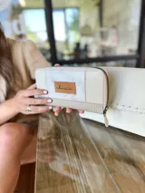 Billetera grande con cierre metálico, color beige claro, con paneles texturizados y un parche de cuero marrón con la marca escrita en cursiva.
