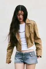 Trench corto de cuello solapa y manga larga, con diseño cruzado y cierre frontal con botones. Presenta bajo acabado en elástico y trabillas en los hombros.
