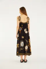 Vestido largo negro de silueta fluida, con finos breteles y detalle fruncido en la parte superior. Presenta un estampado exclusivo con elementos artísticos, palmeras, cisnes y otros motivos.
