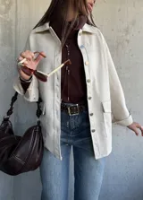 Chaqueta tipo sobrecamisa color beige, con cierre frontal de botones metálicos, cuello clásico y bolsillos frontales. Presenta un diseño de corte recto y mangas largas con puños abotonados.