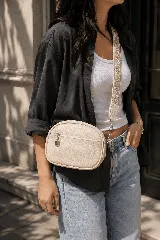 Bandolera beige de diseño ovalado, con bolsillo frontal con cierre y tirador metálico. Cuenta con una correa ajustable y desmontable con diseño étnico bordado.