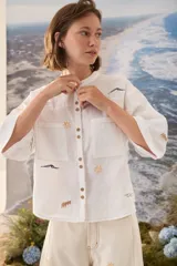 Camisa de lino blanca con cuello mao, mangas cortas y cierre frontal de botones. Presenta delicados bordados multicolores distribuidos por la prenda y bolsillos frontales en el pecho.