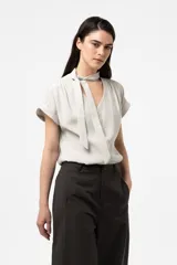 Camisa gris de corte relajado, con escote en V y lazo largo incorporado en el cuello. Mangas cortas con vuelta y frunce sutil en los hombros.