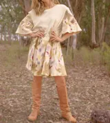 Vestido corto acampanado color crudo, con estampado artesanal de hojas en tonos marrones, violetas y amarillos. Tiene cuello redondo y mangas cortas amplias.