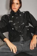 Camisa de cuero ovino color negro, con mangas abullonadas, cuello clásico y cierre frontal con botones metálicos. Presenta detalles bordados con pedrería en forma de estrellas y la frase 'Stars of the night'.