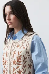 Chaleco tejido color marrón con bordados artesanales de motivos florales y geométricos en color crema. Presenta cuello redondo, cierre frontal con botones y diseño sin mangas.