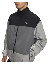 Campera deportiva Under Armour con cierre frontal completo, diseño de bloques de color en tonos negro y gris. Presenta cuello alto, bolsillos laterales con cierre y puños elásticos. Incluye tecnología UA Storm que repele el agua.