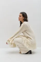 Sweater de lana merino color beige, con silueta amplia y relajada. Presenta mangas largas con volumen, terminaciones con volados de lino y flecos en los puños y en el ruedo, además de detalles trenzados a lo largo de las mangas.