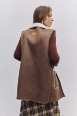 Chaleco largo de gamuza marrón con interior y bordes de corderito sintético blanco. Presenta bolsillos frontales amplios con ribetes de corderito.