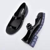 Mocasín estilo Mary Jane de la marca Melissa, modelo June Ad, confeccionado en material sintético negro con acabado brillante. Cuenta con tira ajustable en el empeine con hebilla gruesa, puntera reforzada en tono mate y suela translúcida de goma con diseño chunky.