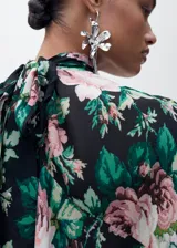 Blusa de gasa con estampado floral en tonos rosa, verde y negro. Tiene cuello alto, mangas largas con volantes y cierre de lazo en la parte posterior.