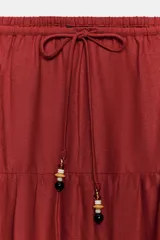 Falda midi color rojo oscuro con diseño de paneles fruncidos, cintura elástica con cordón ajustable y abalorios decorativos.